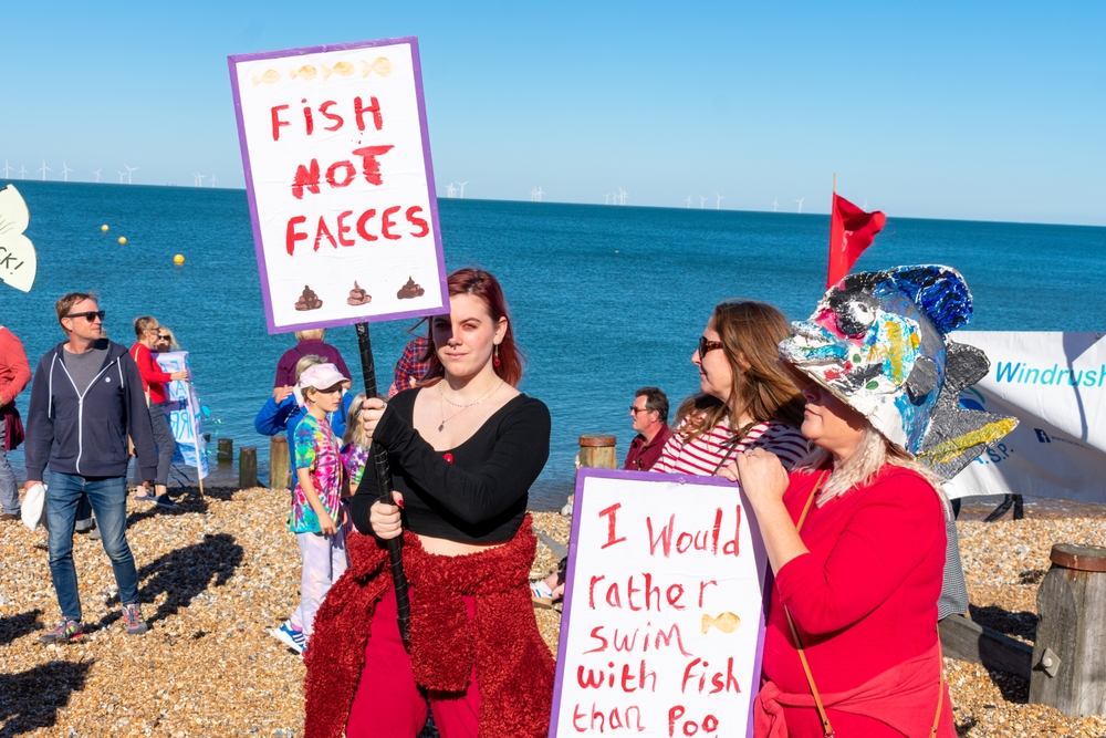Two sewage protesters can be seen with signs at a protest reading "FISH NOT FAECES" and "I would rather swim with fish than poo".