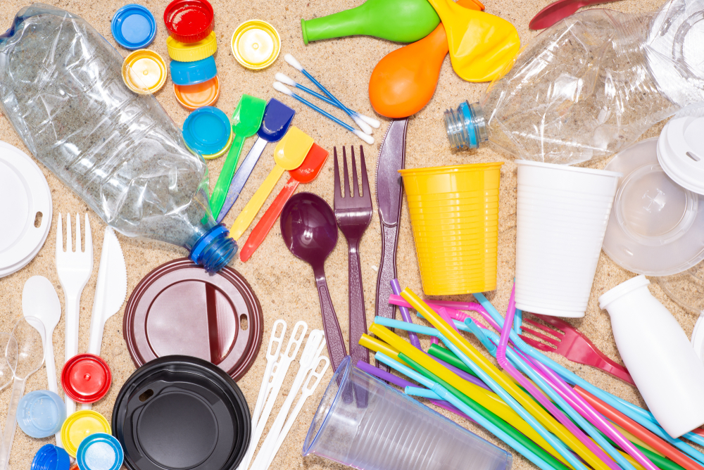 Disposable single-use plastic objects such as bottles, cups, forks, spoons and drinking straws that cause pollution of the environment, especially oceans. Top view on sand