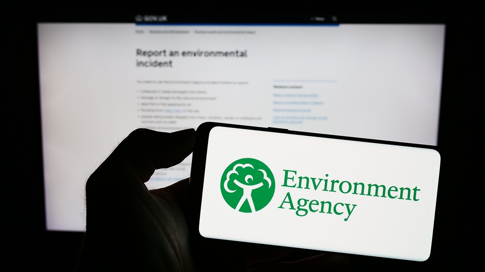Person holding mobile phone with logo of British public body Environment Agency (EA) on screen in front of web page. Focus on phone display.