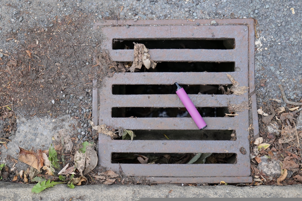 purple single-use vape has been discarded and left lying on a metal water drain cover.